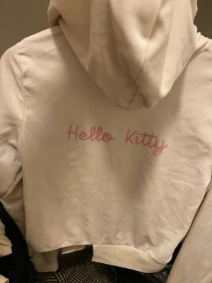 Vit Hello Kitty hoodie från Gina Tricot - Gjort själv, men kofta från Gina men tryckte hello kitty själv Säljer en vit hoodie med dragkedja från Gina Tricot. På ryggen står det 'Hello Kitty' i rosa text. Hoodien har huva med dragsko och två fickor framtill. Perfekt för dig som gillar söta och avslappnade plagg.