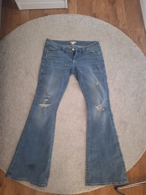 Blå utsvängda jeans med slitningar - Säljer ett par ljusblå utsvängda jeans med slitningar på knäna och broderade detaljer på bakfickorna. Jeansen har klassisk femficksdesign och låg midja. Perfekta för dig som gillar en retro och avslappnad stil. Använda en gång.