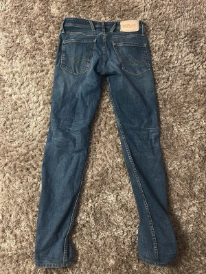 Blå slim fit jeans från Replay - Snygga blå jeans från Replay, modell Anbass med slim fit-passform. Jeansen har klassisk femficksdesign, tydliga sömmar och är tillverkade i ett hållbart material från Sustainable Lab. Perfekta för en trendig och avslappnad look. 27/32