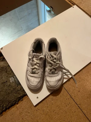 Vita Nike Air Force 1 sneakers - Klassiska vita Nike Air Force 1 sneakers med lågt skaft och ikonisk swoosh på sidorna. Skorna har perforerad tå för ventilation, tjock sula och är tillverkade i läder med textildetaljer. Perfekta för en clean och stilren look.