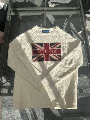 Morris Knit Sweater UK Flag Beige/Vit - Clean Morris knit sweater med stor UK flagga och embroidered logo framtill. Beige/vit colorway med riktig preppy, old money och vintage vibe. Perfekt för dig som gillar british style, y2k, streetwear och casual fits.  Stickad i 100% bomull med soft knitwear feel och skön passform. Enkel att styla med baggy jeans, cargos eller chinos för en retro och classic look. En tidlös vintage inspired sweater som passar året runt.  • Size M • Bra skick • 100% cotton • Preppy / old money aesthetic • Vintage 