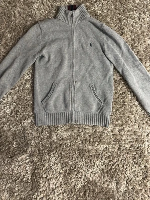 Grå stickad ziptröja Polo Ralph Lauren - Snygg grå stickad ziptröja från Polo Ralph Lauren med hög krage och två fickor framtill. Tröjan har ribbade muddar och nederkant samt en liten broderad logga på bröstet. Perfekt för lager på lager och chill stil.