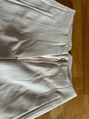 Amisu Beiga kostymbyxor med pressveck - Stilrena beiga kostymbyxor med hög midja och raka ben. Byxorna har pressveck framtill, diskreta sidfickor och dold dragkedja med hake. Perfekta för en dressad look och passar till många olika outfits.