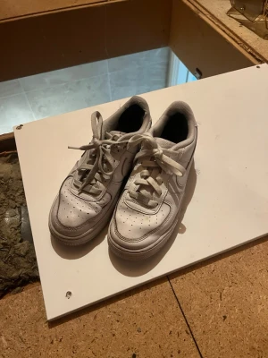 Vita Nike Air Force 1 sneakers - Klassiska vita Nike Air Force 1 sneakers med lågt skaft och perforerad tåbox för extra ventilation. Skorna har en chunky sula, vita skosnören och diskret Nike-logga på sidan. Tillverkade i läder med textilfoder för skön komfort.