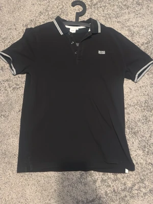 Svart polotopp från BOSS - Klassisk svart polotopp från BOSS med vit logga på bröstet och vita detaljer på krage och ärmslut. Kortärmad modell med knappar framtill och stilren design. Perfekt för en clean och snygg look.
