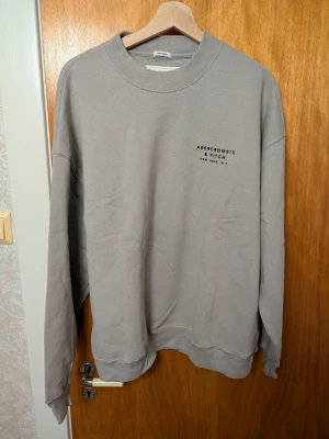 Oversized grå sweatshirt Abercrombie - Grå oversized sweatshirt från Abercrombie & Fitch med rund hals och diskret logga på bröstet. Tillverkad i mjuk fleece för extra komfort och har långa ärmar. Perfekt för en avslappnad och trendig look.
