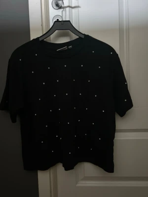Svart t-shirt med pärldetaljer - Svart t-shirt med korta ärmar och små pärlor utspridda över hela framsidan. Enkel och stilren design som passar perfekt till jeans eller kjol. Mjuk och bekväm bomullskvalitet.
