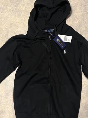 Svart hoodie från Polo Ralph Lauren - Svart hoodie från Polo Ralph Lauren med dragkedja framtill, klassisk huva och fickor. Ikoniska vita loggan broderad på bröstet. Mjuk bomullskvalitet och stilren design som passar perfekt till jeans eller joggers.