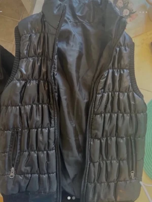 Svart glansig dunväst med dragkedjor - Snygg svart dunväst med glansig finish och quiltat mönster. Västen har hög krage, två fickor med dragkedja och ribbade muddar vid ärmhålen. Perfekt för lager-på-lager och ger en cool streetstyle vibe.