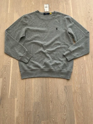 Grå sweatshirt från Polo Ralph Lauren - Klassisk grå sweatshirt från Polo Ralph Lauren med rund halsringning och broderad marinblå logga på bröstet. Tröjan har långa ärmar och ribbade muddar vid ärmslut och nederkant. Perfekt för en stilren och avslappnad look.