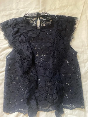 Mörkblå spetsblus från Zara - Supersnygg och gullig  mörkblå blus i spets från Zara med hög hals och knäppning bak.  Blusen är ärmlös och har fina volangdetaljer framtill samt vackert blommönster i spetsen. Har använt den en gång men säljer den för den kommer aldrig till användning.