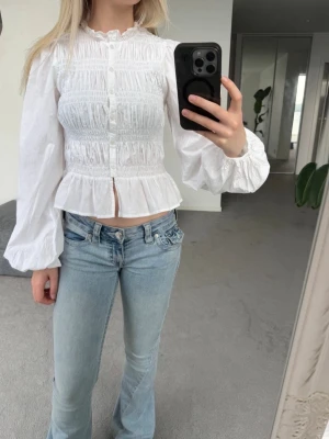 Vit blus med smock - Supersöt vit blus med smockad överdel, volang nertill och knappar framtill. Blusen har långa puffärmar med resår vid ärmslut och en romantisk känsla. Perfekt att styla med jeans för en trendig look.