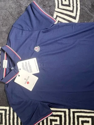 Marinblå Moncler pikétröja - Snygg marinblå pikétröja från Moncler med klassisk krage och knäppning framtill. Tröjan har röda och vita ränder på krage och ärmslut samt Moncler-logga på bröstet. Perfekt för en stilren och sportig look.