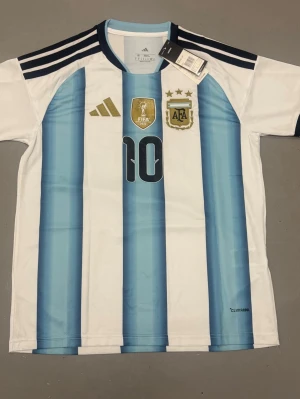 Argentinas vm tröja messi - Snygg Argentina matchtröja från Adidas med Messi och nummer 10 på ryggen. Tröjan har klassiska blå och vita ränder, svarta detaljer och guldiga loggor på bröstet. Tillverkad i lätt och ventilerande material, perfekt för fotbollsträning eller match.