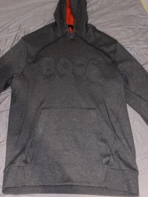 Mörkgrå hoodie från BOSS - Mörkgrå hoodie från BOSS med broderad logga på bröstet. Hoodien har svart snörning, känguruficka och en kontrasterande orange insida i huvan. Perfekt för en chill och stilren look.