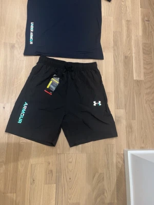 Under Armour svart träningsset - Snyggt träningsset från Under Armour med svart t-shirt och matchande shorts. T-shirten har logga och text i vitt och turkost på sidan. Shortsen har elastisk midja, dragsko, logga och text i vitt och turkost samt reflekterande detaljer bak. Perfekt för gym eller för att vara ute och ta det lungt 