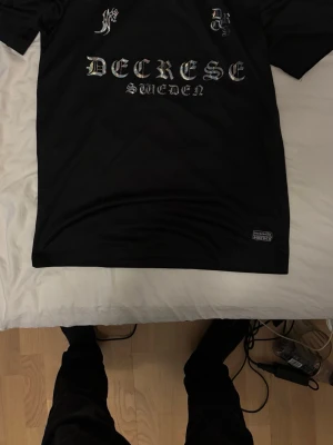Svart t-shirt Decrese Sweden - Svart t-shirt från Decrese Sweden med holografiskt tryck i gotisk stil på bröstet. T-shirten har rund hals och rak passform. Perfekt för dig som gillar streetwear och unika detaljer.