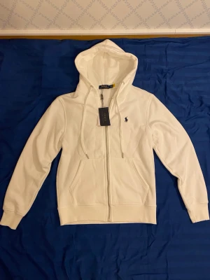 Vit hoodie från Polo Ralph Lauren - Snygg vit hoodie från Polo Ralph Lauren med dragkedja framtill och klassisk huva. Broderad logga på bröstet och två fickor framtill. Tillverkad i mjukt bomullsmaterial som är skönt att ha på sig. Perfekt för en chill och stilren look.
