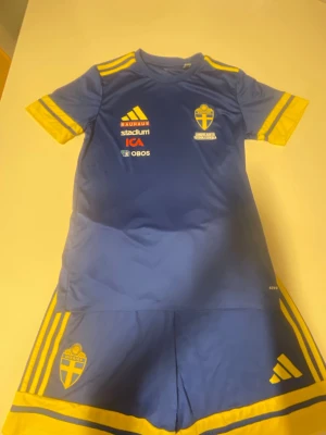 Svenska landslaget fotbollskit blå/gul - Säljer ett blått och gult fotbollskit från Adidas med Svenska landslagets logga. Setet består av en t-shirt och shorts med gula detaljer och sponsormärken på bröstet. Perfekt för träning eller match. Materialet är lätt och andas.