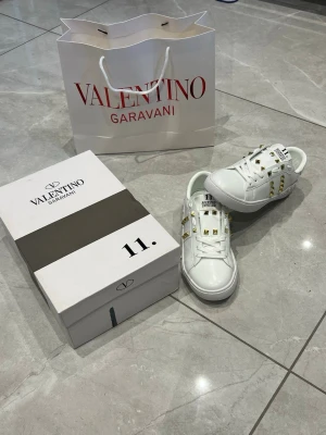 Valentino  vita sneakers - Snygga vita sneakers från Valentino Garavani med ikoniska guldiga nitar på sidorna och ovandelen. Skorna har klassisk låg modell, vita skosnören och detaljerad logga på plösen. Tillverkade i läder för en lyxig känsla och stilren look.