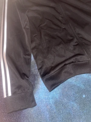 Svart träningsjacka med vita ränder - Snygg svart träningsjacka med vita ränder längs ärmarna. Jackan har muddar vid ärmslut och nederkant samt praktiska fickor på sidorna. Tillverkad i ett glansigt, sportigt material som passar perfekt till träning eller chill.