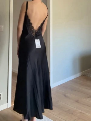 Svart satinklänning med öppen rygg och spetsdetaljer - Så fin klänning, aldrig använd med prislappen kvar. Går att justera banden beroende på hur djupt man vill att öppningen ska gå/hur den ska sitta vid bröstkorgen🥰 Det finns många fler bilder på deras hemsida men jag får bara ta med egna bilder. Sök på Design by si Minnie satin dress, så kommer den upp❤️Storleken är XS\S och tycker det stämmer bra överens med hur klänningen passar🥰
