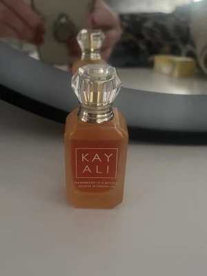 Kayali Orange Blossom parfym - Kayali parfymflaska i orange glas med elegant, kristalliknande lock och silvriga detaljer. Doften heter Marrakesh In A Bottle Orange Blossom 24 och flaskan har en modern, stilren design som sticker ut på sminkbordet.