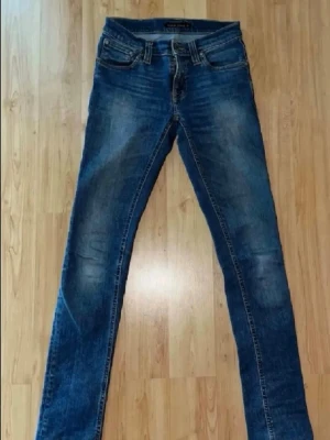 Blå slim fit jeans från Nudie Jeans - Säljer ett par klassiska blå slim fit jeans från Nudie Jeans med snygga slitningar och kontrastsömmar. Jeansen har fem fickor, normalhög midja och är tillverkade i bomull med stretch för extra komfort. Perfekta för en avslappnad och trendig look.