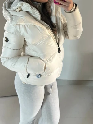 Vit pufferjacka med huva - Snygg vit pufferjacka med dragkedja, huva och detaljer i svart. Jackan har två fickor framtill och justerbar nederkant. Perfekt för kalla dagar och har en modern, puffig look. Märke syns på ärmen.