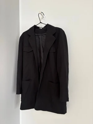 Svart lång kavaj med fickor - Stilren svart kavaj med klassisk krage och två stora framfickor. Kavajen har en rak passform och är helfodrad för extra komfort. Perfekt för en trendig och avslappnad look. Inga synliga knappar framtill och diskreta bröstfickor med lock.