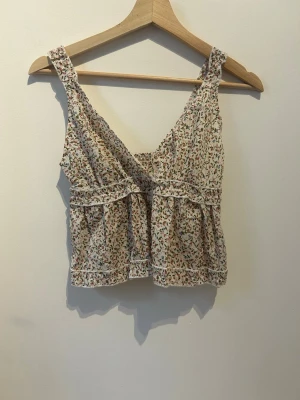 Blommig linnetopp med volang - Söt ljus linnetopp med blommigt mönster i rosa, grönt och brunt. Toppen har breda axelband, v-ringning och volangdetaljer framtill. Smockad rygg för skön passform och luftig känsla. Perfekt för varma dagar och enkel att styla.