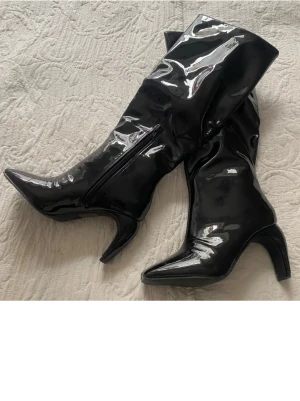 lackstövlar - Säljer ett par svarta höga stövlar i glansigt lackmaterial med spetsig tå och markerad klack. Stövlarna har dragkedja på insidan och en cool, trendig look som sticker ut. Perfekta för dig som vill ha statement boots till din outfit.