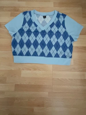 Ny.Blå argyle kortärmad tröja - Snygg ljusblå kortärmad tröja med klassiskt argylemönster i blått och vitt. Tröjan har v-ringning och ribbade kanter. Perfekt för dig som gillar preppy stil och vill sticka ut lite extra. Storlek XL men förminskar som L