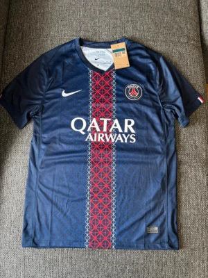 PSG matchtröja blå Nike 25/26 home - Säljer en blå Paris Saint-Germain matchtröja från Nike med Qatar Airways-tryck på bröstet. Tröjan har ett rött och vitt mönster längs mitten, PSG-logga och Nike Swoosh på framsidan. Tillverkad i lätt och ventilerande material, perfekt för fotboll.