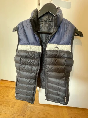 J.Lindeberg blå och svart dunväst - Dunväst från J.Lindeberg. Går att vika in och ut, svart på ena sidan och blå/svart på andra. Är en storlek M, men är liten. Sitter mer som en S