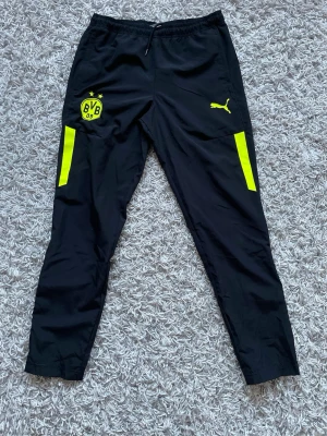 Svarta BVB träningsbyxor från Puma - Snygga svarta träningsbyxor från Puma med Borussia Dortmunds gula klubbmärke och logga på vänster ben. Byxorna har gula detaljer längs benen och elastisk midja med dragsko. Perfekta för fotbollsträning eller gymmet.