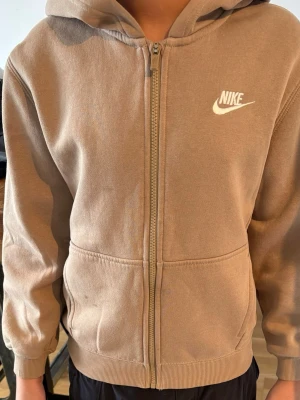 Beige zip hoodie från Nike - Säljer en beige hoodie från Nike med dragkedja framtill och vit Nike-logga på bröstet. Hoodien har huva, ribbade muddar och två fickor fram. Tillverkad i mjukt bomullsmaterial som är skönt att ha på sig.