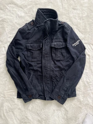Abercrombie & Fitch Field Jacket M - Snygg svart jacka från Abercrombie & Fitch med hög krage, fyra framfickor och knappdetaljer på axlarna. Jackan har broderad logga på ärmen och stängs med dragkedja och knappar. Tillverkad i bomull för en skön och avslappnad look.