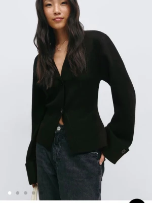 Svart kofta från Gina Tricot - Svart kofta med v-ringning och knäppning framtill från Gina. Modellen har långa ärmar med breda muddar och en något croppad passform. Har ägt den i ungefär ett halvår och den är använd en handfull gånger. 