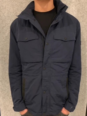 Mörkblå field jacket från J.Lindeberg - Stilren mörkblå vindjacka från J.Lindeberg med hög, Tillverkad i ett lätt och slitstarkt material som står emot vind. Lite tjockare material så perfekt under dagens väder och hösten också. Storlek M, skriv gärna cid intresse!