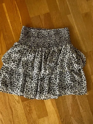 Leopardmönstrad volangkjol - Supersöt kort kjol med leopardmönster i svart och vitt. Kjolen har bred smock i midjan och dubbla volanger som ger en fluffig och trendig look. Perfekt för dig som vill sticka ut lite extra. Materialet känns lätt och luftigt.