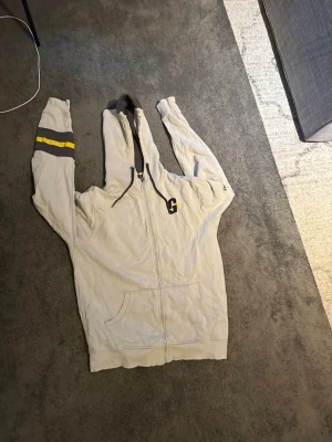 Grå Gymgrossisten hoodie med dragkedja - Säljer en grå hoodie från Gymgrossisten med dragkedja framtill och huva med snören. Stora gula och svarta detaljer, bland annat logga på bröstet och text längs ryggen. Två fickor fram och coola ränder på ena ärmen. Perfekt för gymmet eller chill.