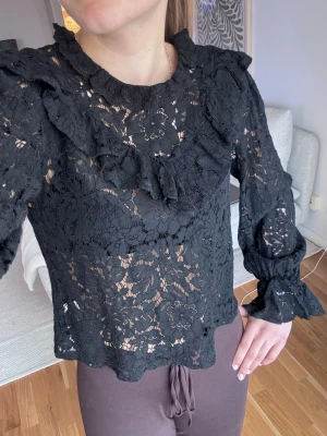 Svart spetsblus med volangdetaljer - Trendig svart blus i genombruten spets med volangdetaljer längs ringningen och axlarna. Blusen har långa ärmar med resår vid ärmslut och en feminin, romantisk look. Perfekt att styla med både jeans och kjol för en cool outfit.