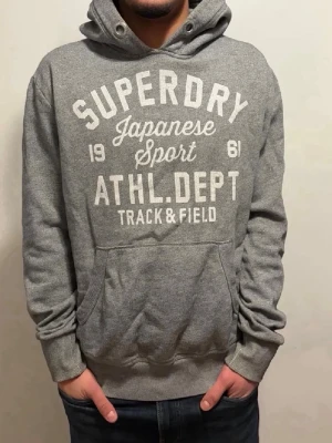Extremt sällsynt Grå Superdry hoodie med tryck - Snygg grå hoodie från Superdry med stort vitt tryck på bröstet där det står 'Superdry Japanese Sport ATHL. DEPT Track & Field'. Klassisk känguruficka framtill och justerbar huva med dragsko. Perfekt för en avslappnad streetwear-look.