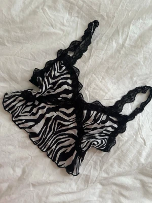 Zebramönstrad spetstop - Säljer en snygg zebramönstrad top med svarta spetsdetaljer och volangkant. Toppen har breda axelband i spets och är gjord i ett tunt, genomskinligt material. Perfekt för dig som gillar djurmönster och vill ha något unikt i garderoben.