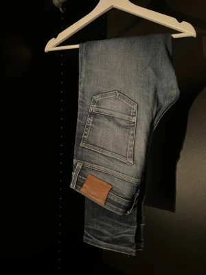 Blå slitna jeans från Jack & Jones - Snygga blå slim fit jeans från Jack & Jones med slitningar och ljusa partier. Klassisk femficksmodell med knappgylf och läderpatch bak i midjan. Perfekta för en avslappnad och trendig look.