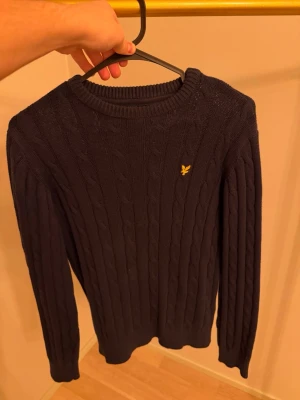 Mörkblå kabelstickad tröja Lyle & Scott - Säljer en mörkblå kabelstickad tröja från Lyle & Scott med rund halsringning och gul broderad logga på bröstet. Tröjan har långa ärmar och ribbade muddar vid ärmslut och nederkant. Perfekt för lager på lager och klassisk stil.