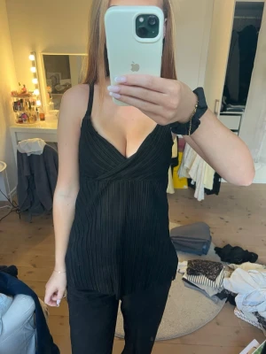 Svart plisserad linnetopp - Snygg svart linnetopp med smala axelband och v-ringad omlottdesign. Toppen har ett plisserat tyg som ger en cool struktur och faller fint. Perfekt att styla med jeans eller kjol för en trendig look. Banden är justerbara!