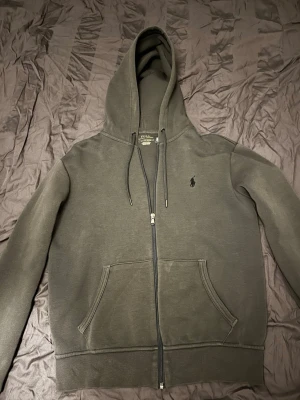 Mörkgrå hoodie från Polo Ralph Lauren - Mörkgrå hoodie med dragkedja från Polo Ralph Lauren. Klassisk design med huva och dragsnören, samt känguruficka framtill. Liten broderad logga på bröstet. Perfekt för en avslappnad och stilren look. Storlek S