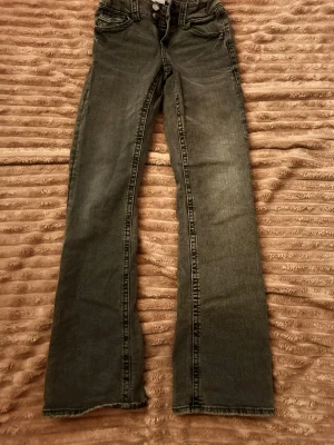 Grå bootcut jeans med låg midja - Säljer ett par grå bootcut jeans med låg midja och klassiska fem fickor. Jeansen har en lätt tvättad look och raka ben med något utsvängd passform. Perfekta till sneakers eller boots för en avslappnad stil.